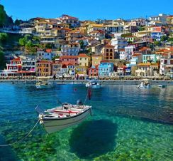 A small white boat floats on clear turquoise water near a coastal town with colorful houses stacked on a hillside under a blue sky.