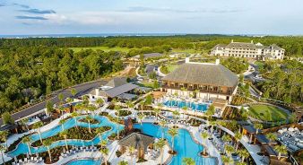 Aerial view of Camp Creek Inn - a luxury resort with multiple outdoor swimming pools, lounge chairs, palm trees, large buildings, and green landscape in the background.