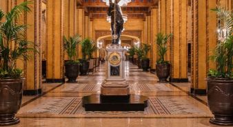 The Roosevelt New Orleans grand hallway with tall potted plants, ornate golden walls, patterned floor, and a prominent statue on a pedestal in the center. Chandeliers hang from the ceiling.