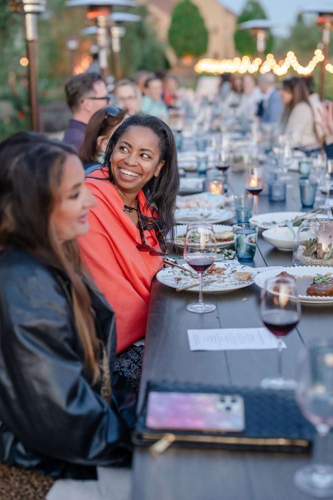 People seated at a long outdoor dinner table with food, drinks, and candles, engaged in conversation. One woman in a red wrap smiles at another guest.