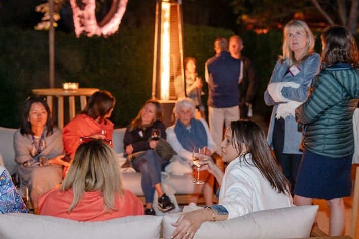 A group of people are seated and standing around an outdoor heater at night, some holding drinks and conversing, with a blurred background of greenery.