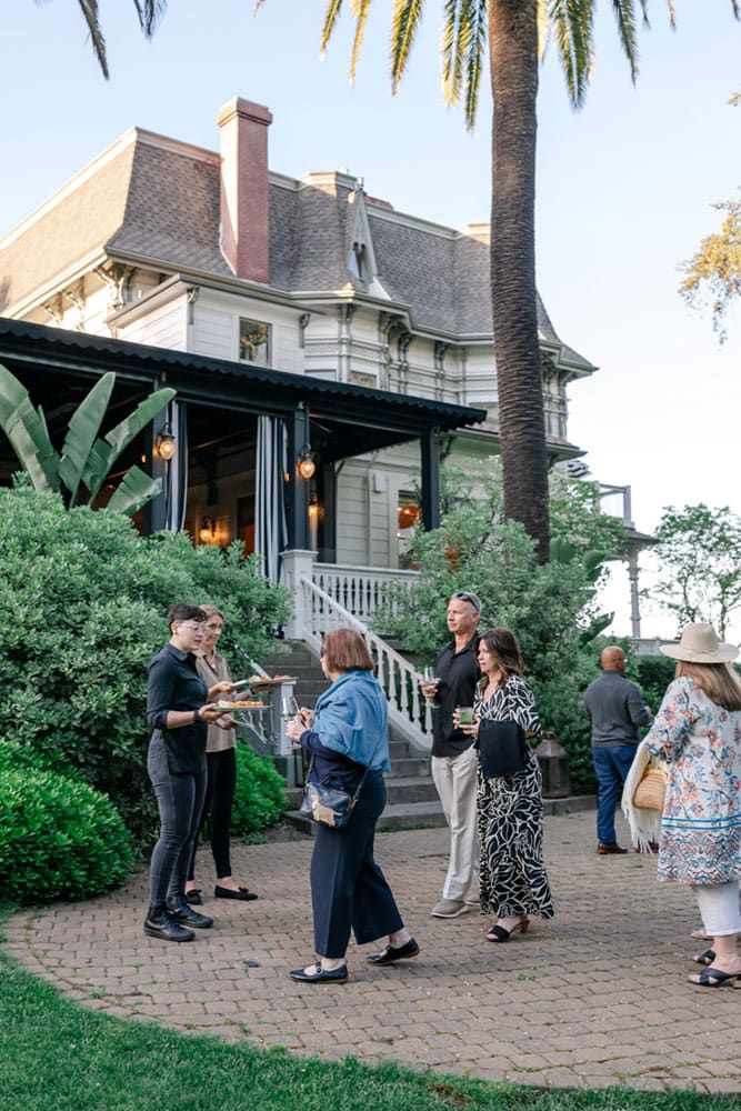 A group of people stand and converse on a brick patio in front of a large house, with two people serving drinks and food on trays.