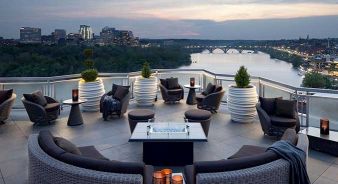 The Watergate Hotel’s Top of the Gate rooftop bar with modern seating, large planters, and a fire pit overlooking the Potomac River and Washington, DC at dusk.