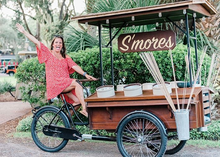 A woman in a red dress sits on a tricycle cart labeled "S'mores," which displays empty bowls and marshmallow roasting sticks. Shrubs and trees are in the background.