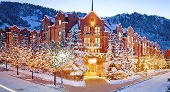 St. Regis Aspen Resort - Aspen, CO. A large red-brick hotel building surrounded by snow-covered trees and mountains, with warm lights glowing at dusk.
