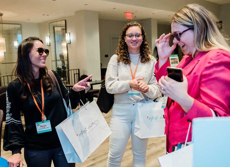 Three women wearing sunglasses stand indoors, holding shopping bags and laughing. One holds a phone; all wear conference name badges.