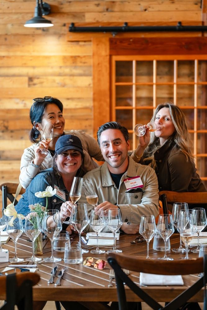 Four people sit at a table with wine glasses, smiling and holding drinks in a cozy, wood-paneled room.