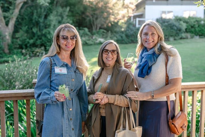 Three women stand on a wooden deck outdoors, smiling at the camera and holding drinks, with greenery and a house visible in the background.