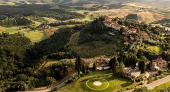 Aerial view of the hilltop luxury resort Castelfalfi surrounded by trees, fields, and rolling hills, with a circular helipad visible in the foreground.