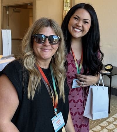 Two women wearing conference badges pose indoors; one is smiling and holding a white Retreats Resources gift bag, while the other is wearing sunglasses.