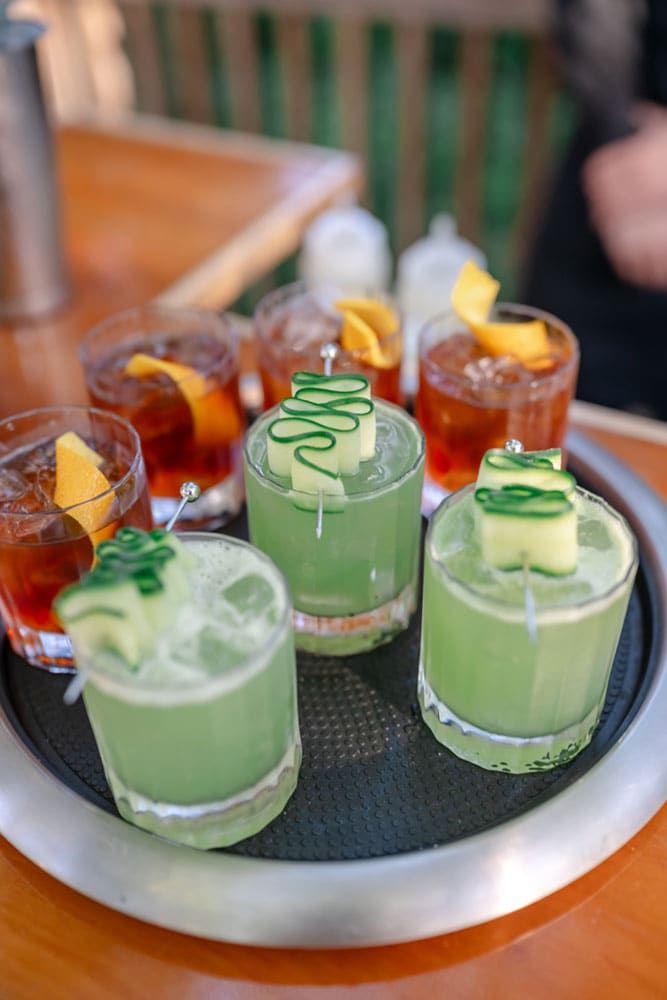 A tray holding six cocktails, with three green drinks garnished with cucumber and three amber drinks garnished with orange peel.