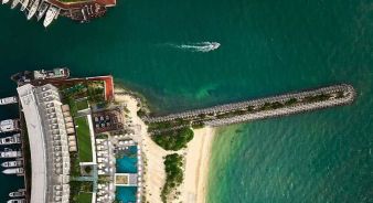Aerial view of SLS Playa Mujeres waterfront resort with pools, a sandy beach, a pier extending into green water, and a boat leaving a wake offshore.
