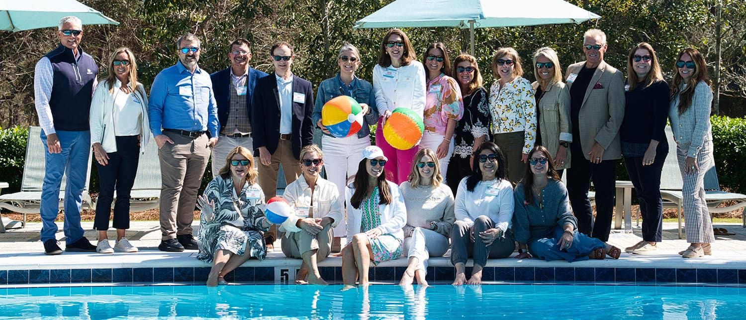 A group of people pose for a photo by a pool, with some seated at the edge and others standing behind them. A few hold beach balls, and everyone is wearing sunglasses.