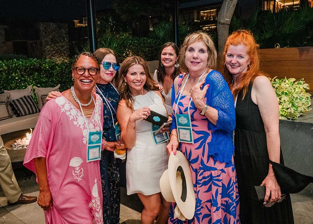 A group of six women smiling and posing together outdoors at a Connoisseurs Circle Conference, some wearing name badges.