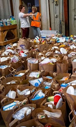 Two people stand by a door while numerous brown paper bags filled with groceries and supplies cover the floor of a warehouse.