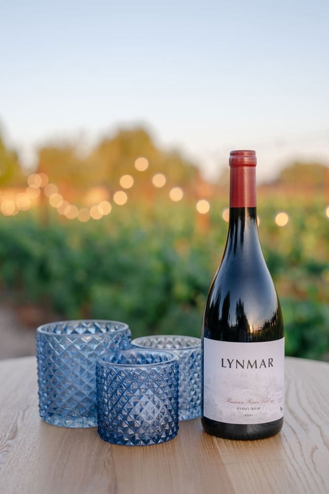 A bottle of Lynmar Pinot Noir and three blue textured glass candle holders sit on a wooden table outdoors with blurred string lights and grapevines in the background.