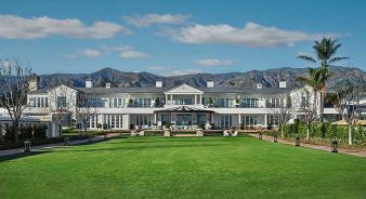 Large white two-story Rosewood Miramar Beach building with a wide lawn in front, outdoor seating area, and mountains in the background under a partly cloudy sky.
