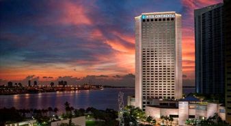 A tall InterContinental hotel building stands by the waterfront at sunset, with a dramatic sky and cityscape in the background.