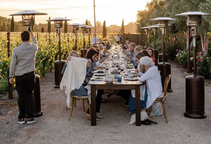 A large group of people dine outdoors at a long table surrounded by patio heaters and greenery during sunset.