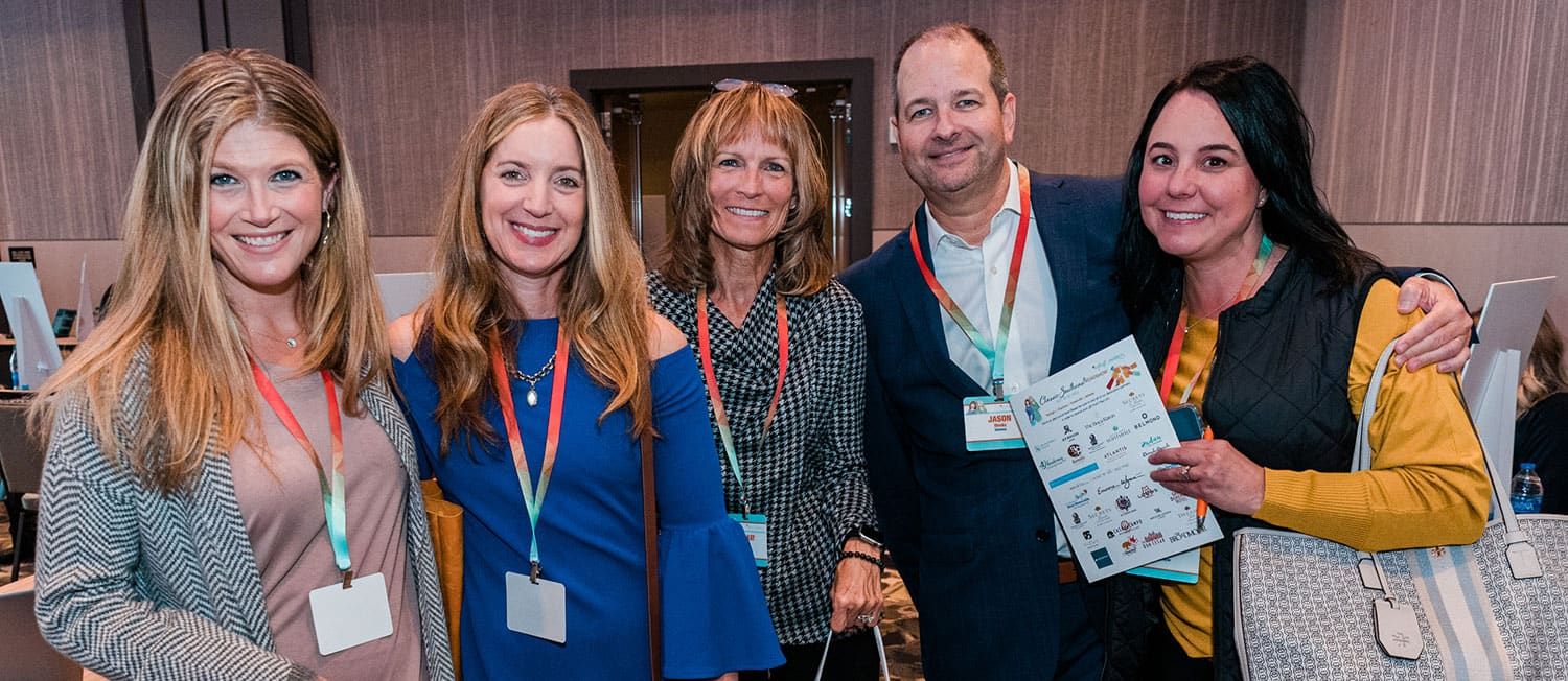 Five smiling people pose together indoors at a Retreats Resources Roadshow event, wearing name badges and lanyards; one person holds a printed sheet with images and text.