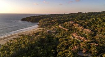 Aerial view of Siari, a Ritz-Carlton Reserve, a coastal resort in Nayarit, Mexico surrounded by dense forest, with buildings scattered along the beach and hillside at sunset.