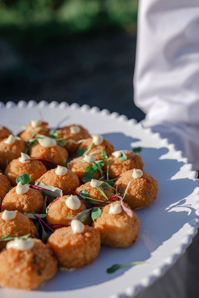 A white plate holds several breaded, bite-sized appetizers topped with dollops of sauce and garnished with microgreens.