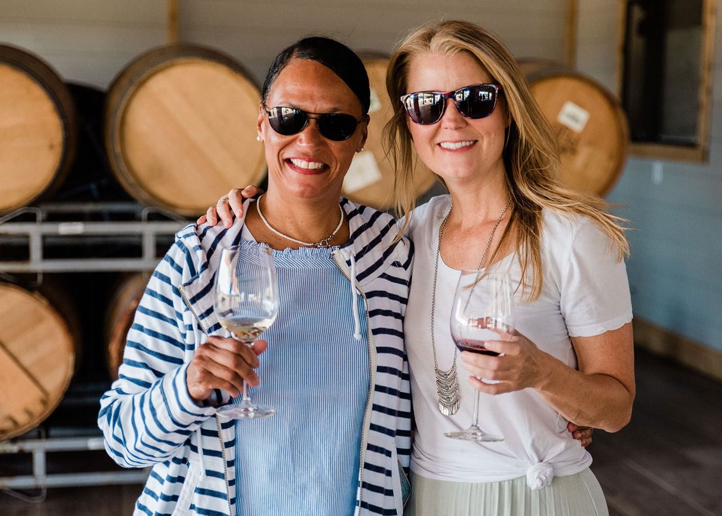 Two women wearing sunglasses stand side by side, smiling and holding wine glasses, with large wooden barrels in the background.