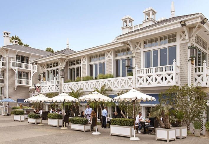 Shutters on the Beach - a two-story seaside restaurant with outdoor seating, white railings, and umbrellas. People are dining and staff are serving at tables on a sunny day.