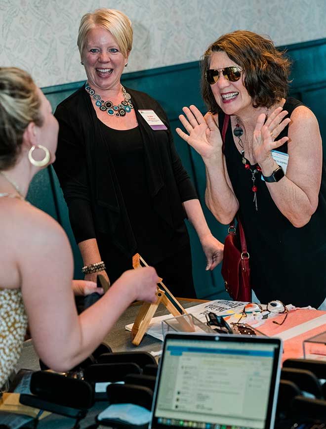 Two women stand smiling and talking with a third woman across a table displaying various Maui Jim gifting items, with a laptop open in the foreground.