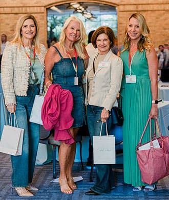 Four women stand together indoors, smiling and holding white goodie bags branded with "Retreats Resources." They wear name tags and are surrounded by tables. A window offers a serene view of trees, setting a perfect scene for the Lone Star State Roadshow destination showcase.
