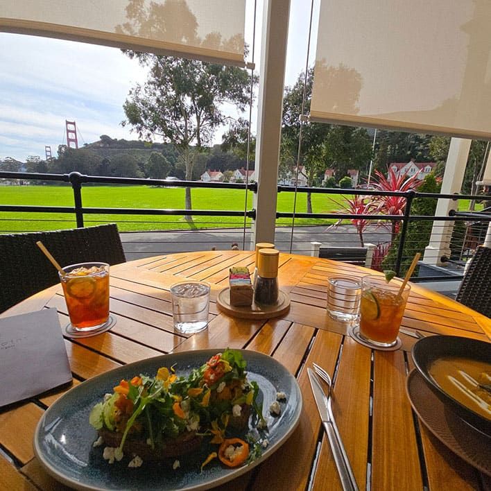 Sula @ Cavallo Point, Sausalito, CA. A wooden patio table set with food and drinks overlooks a green lawn, trees, and the Golden Gate Bridge in the background.