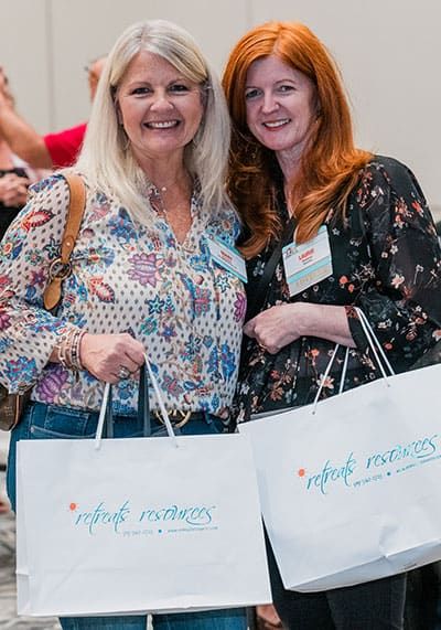 Two women holding white tote bags labeled “retreat resources,” standing side by side and smiling at the camera.