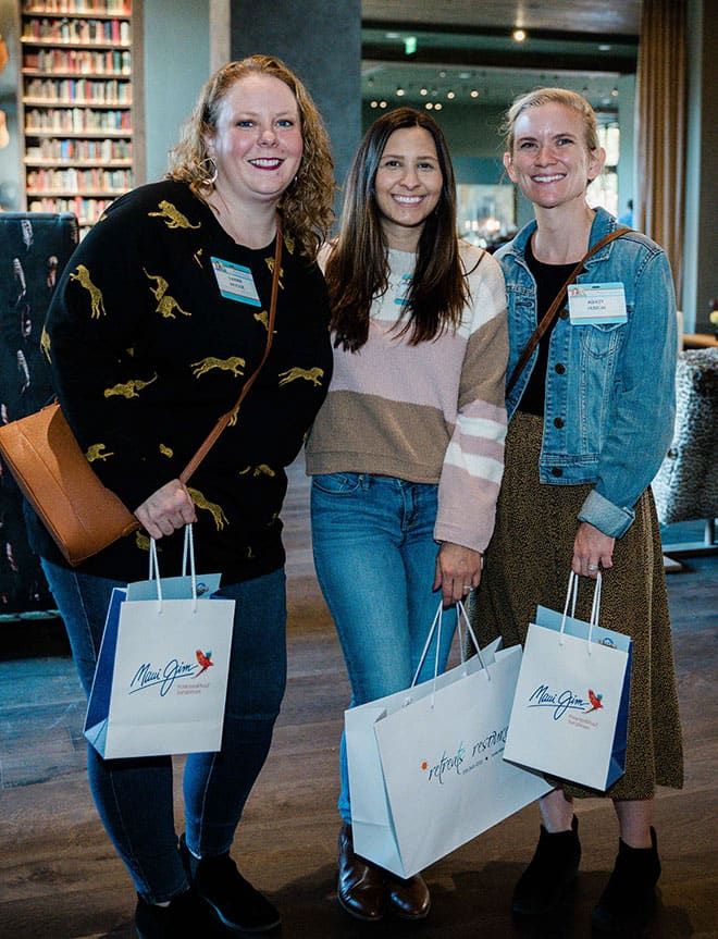 Three women stand indoors, smiling at the camera, holding Retreats Resources and Maui Jim gift bags and wearing name tags. A bookshelf is visible in the background.