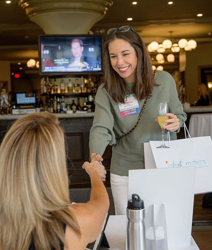 A woman standing and smiling shakes hands with another seated woman at a Retreats Resources Roadshow, holding a drink and a gift bag. A TV is on in the background.