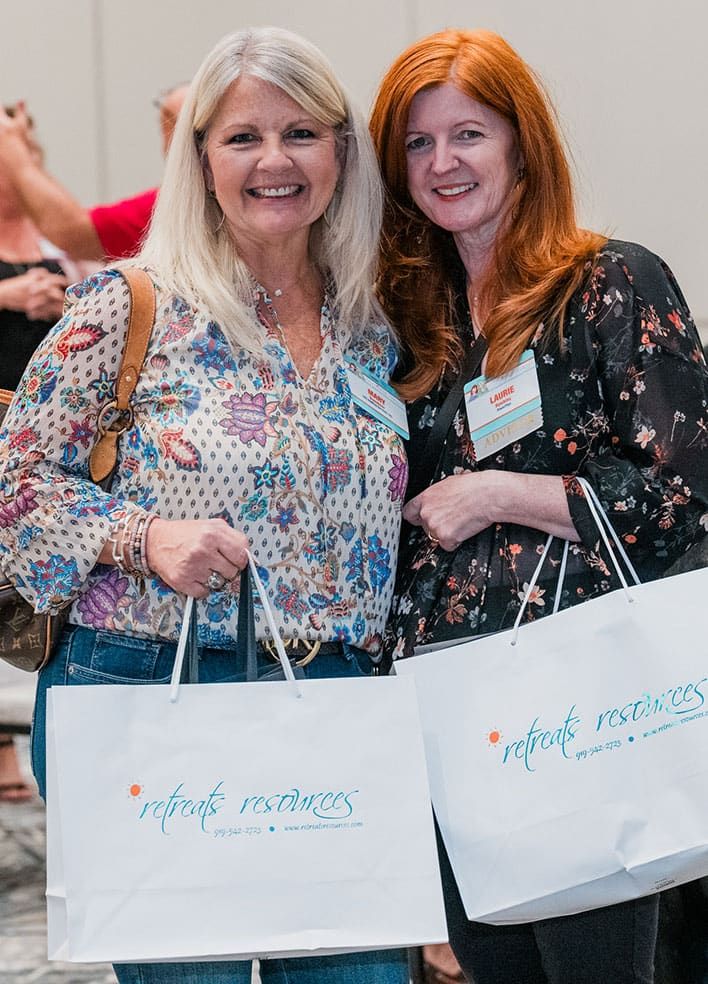 Two women stand indoors, smiling at the camera and holding white "retreat resources" tote bags. Both wear patterned blouses and name badges.