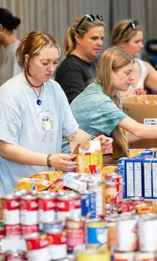 Several people sort and organize canned goods and boxed food items on a table, volunteering with Wine To Water.