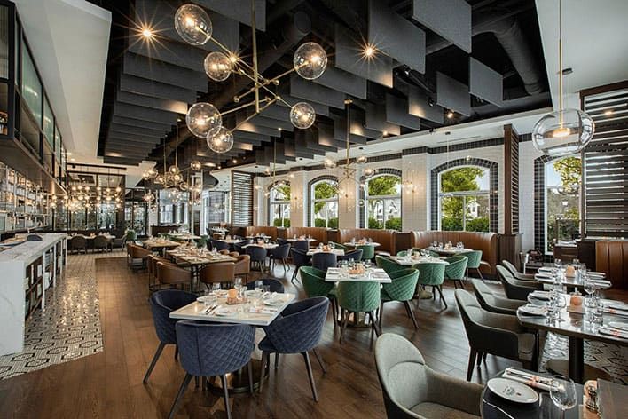 Modern restaurant interior at Orchard Park by David Burke, with large windows, contemporary lighting fixtures, cushioned chairs, set tables, and a mix of dark and natural wood tones.