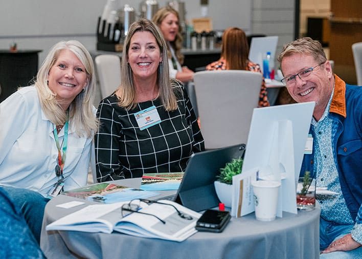 Three adults sit at a round table with papers, a laptop, and drinks, smiling at the camera during a Retreats Resources Roadshow showcase event..