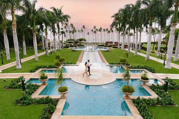 The Conrad Tulum by the ocean featuring an outdoor pool lined with white umbrellas, palm trees, and lounge chairs, with buildings and turquoise water in the background.
