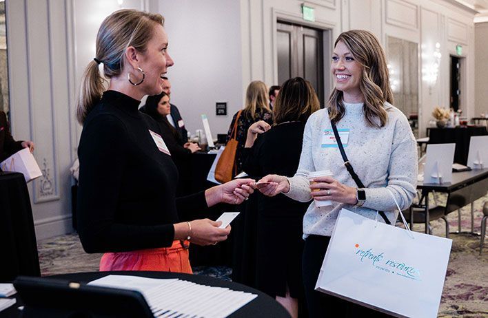 Two women stand and talk at a professional event; one woman hands a business card to the other, who holds a drink and a shopping bag. Several people and tables are in the background.