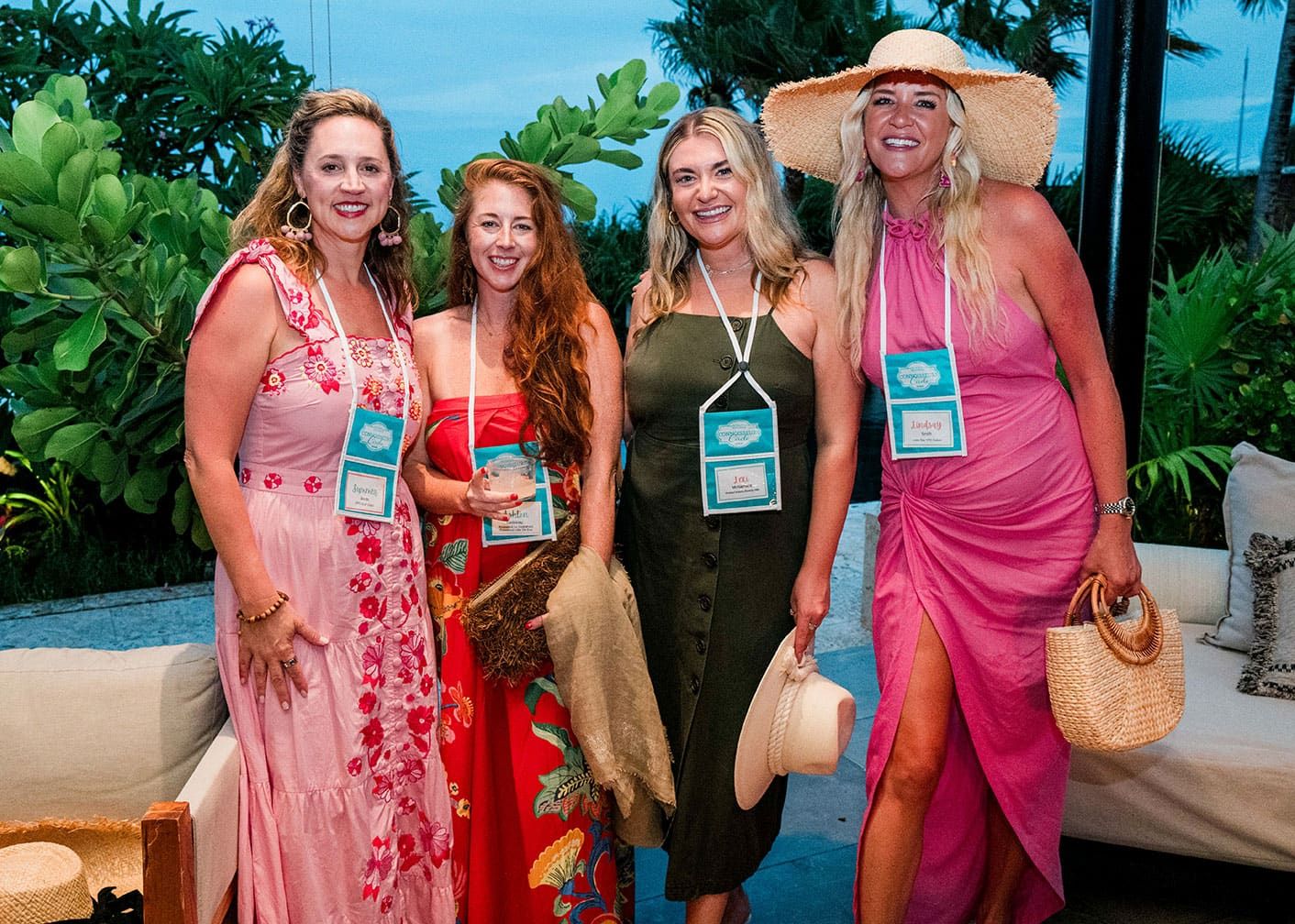 Four women wearing summer dresses and name badges stand together outdoors, smiling, with greenery in the background.