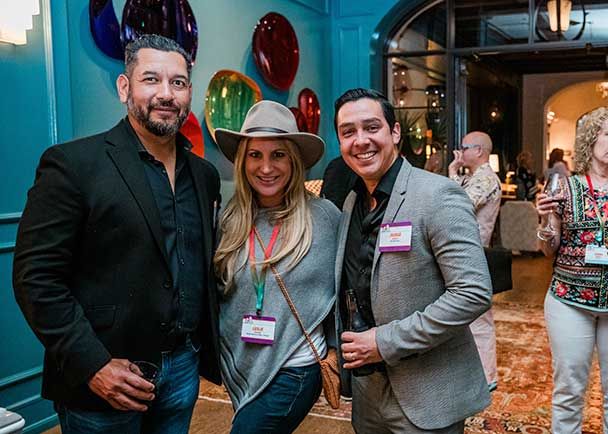 Three people stand together at the Lone Star State Roadshow, smiling at the camera. The woman in the middle sports a stylish hat. Colorful wall decorations are visible in the background, adding to the festive atmosphere of this indoor Retreats Resources event.