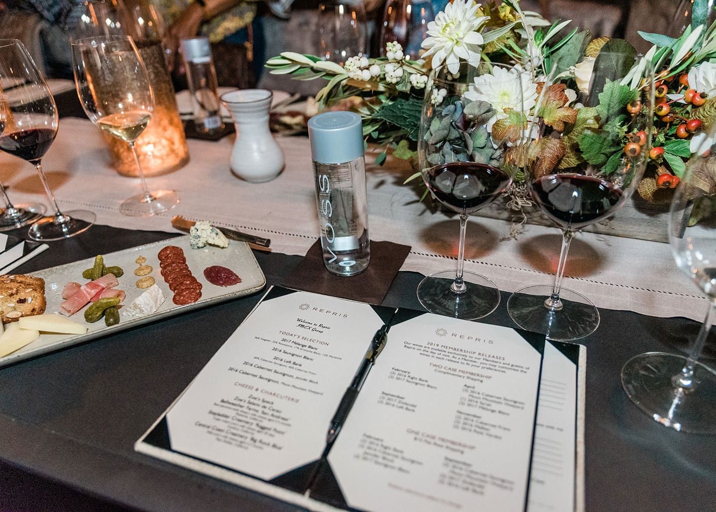 Two wine glasses, a bottle of water, a menu, and a cheese and charcuterie platter are arranged on a table with decorative flowers and candles.
