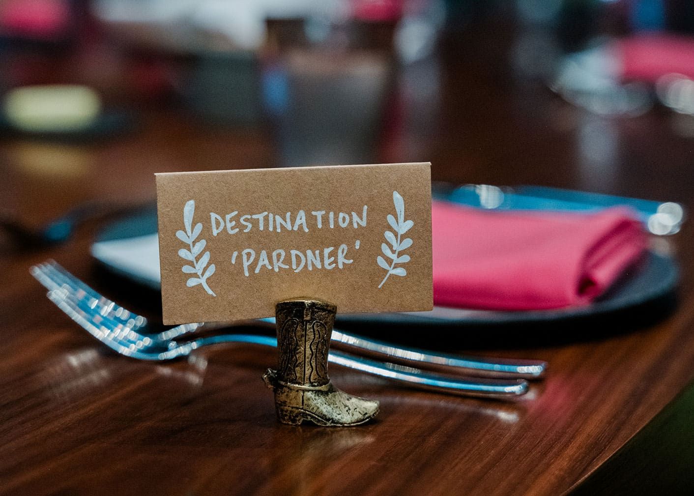 A small card reading "Destination 'Pardner'" is held by a miniature cowboy boot stand on a wooden table set with silverware and pink napkins.