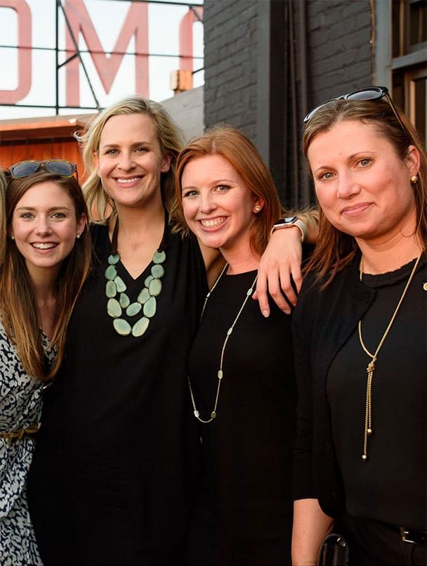 Four women stand closely together outdoors, smiling at the camera. They are dressed in casual and semi-formal attire. A building and partial sign are visible in the background.