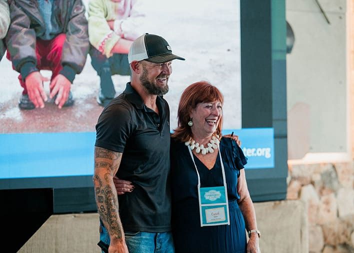 WineToWater Founder Doc Hendley and Retreats Resources Founder & President Carol Owen stand smiling together in front of a large projected image and a blue banner at an indoor event.