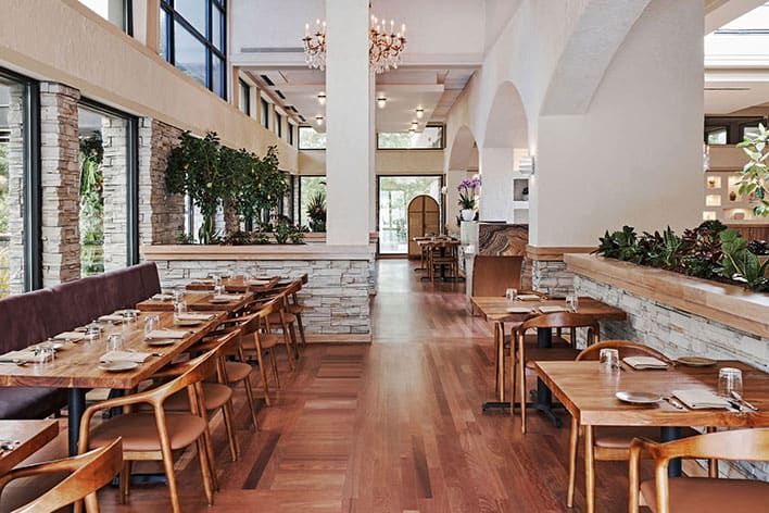 Limani Mediterranean Restaurant in Alpharetta, GA. A contemporary restaurant interior with wooden tables and chairs, set for dining; large windows, stone accents, plants, and natural light fill the space.