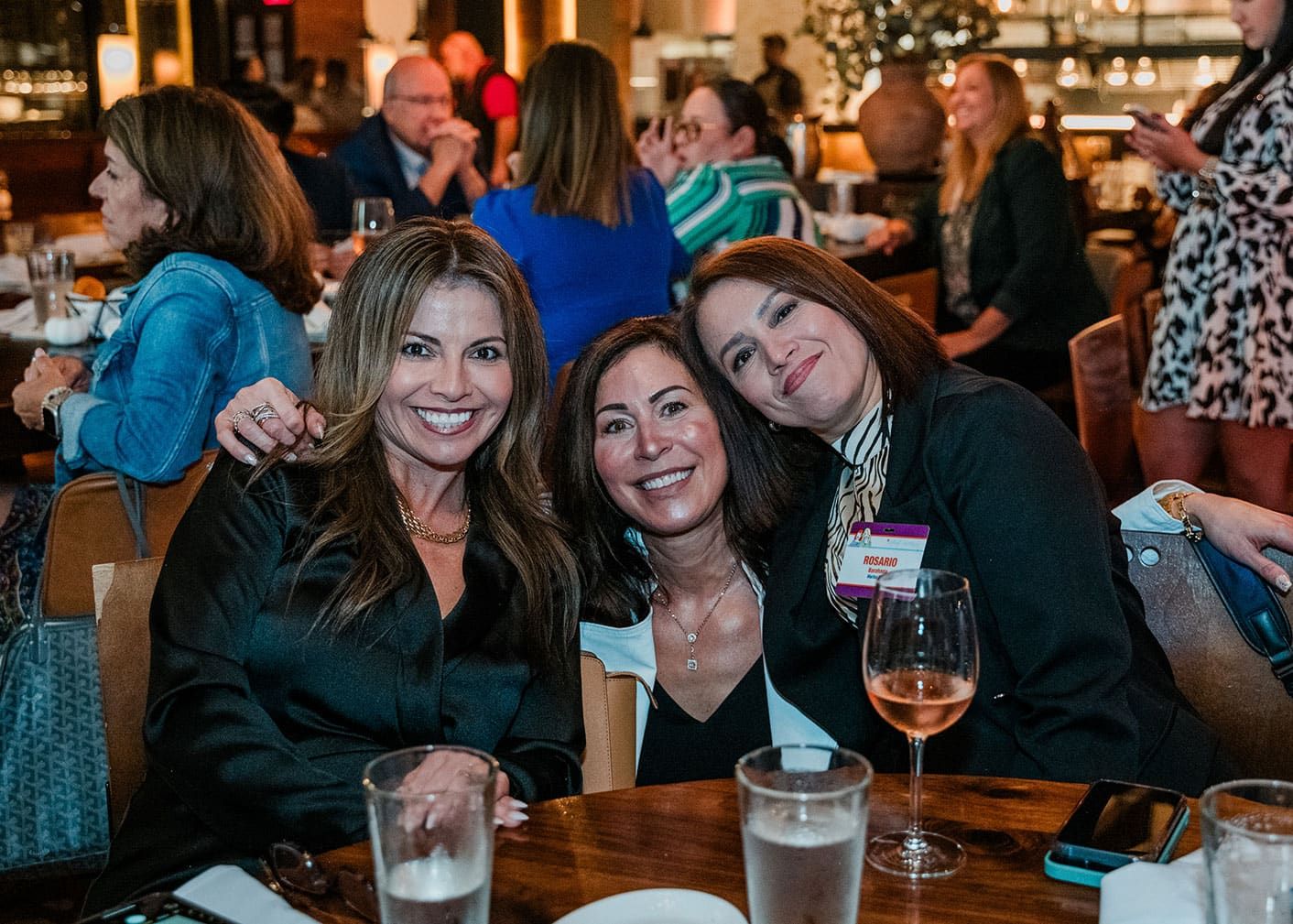 Three women sit closely together and smile at a restaurant table, surrounded by other people dining and socializing in the background.