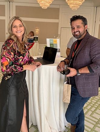 Two people smile for the camera and stand by a round table with a laptop during a Retreats Resources roadshow showcase event. 