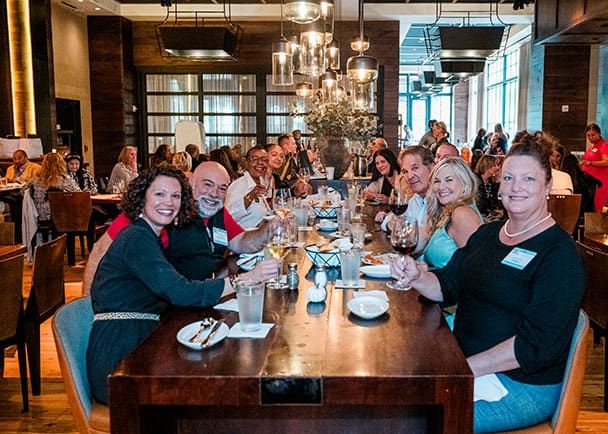 Four women with name tags stand together at a table, smiling and laughing at a Retreats Resources Roadshow. A laptop, coffee cup, and soda can are visible on the table.